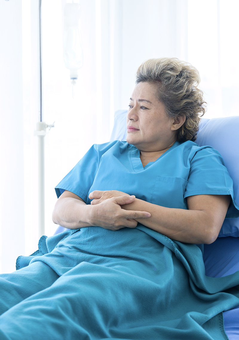 An elderly asian woman lies in a blue hospital gown in a hospital bed.