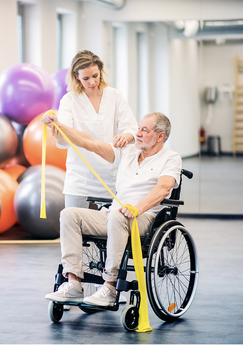 A young white female physiotherapist helps her elderly male patient who uses a wheelchair with his stretching exercises.