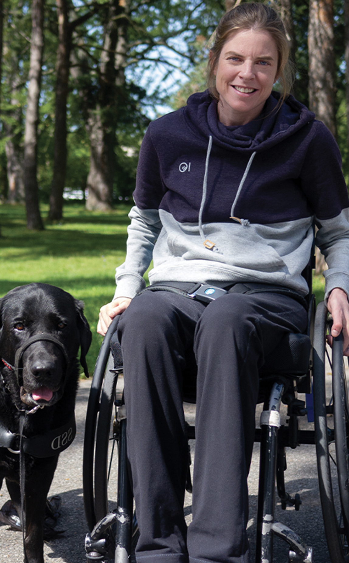 A young white female with blond hair sits in her wheelchair smiling beside her therapy dog.