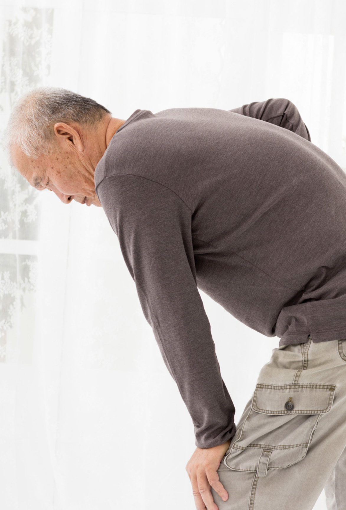 An elderly asian male leans forward and clutches his lower back in pain.